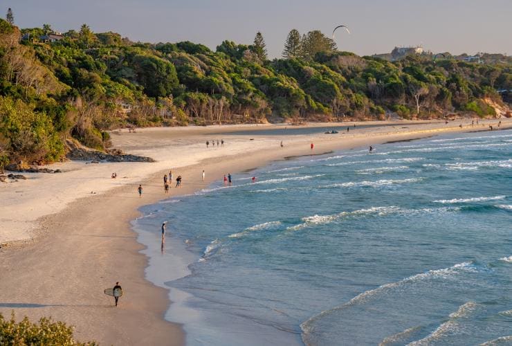 Eine Luftaufnahme des Ozeans und des goldenen Strandabschnitts, der von grünem Buschland gesäumt wird, mit Schwimmern und Surfern am Strand in Byron Bay, New South Wales © Tourism Australia