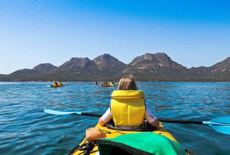 Eine Gruppe von Kajakfahrern auf einer ruhigen blauen Wasserstraße mit Berggipfeln in der Ferne während einer Tour mit Freycinet Adventures, Freycinet National Park, Tasmanien © Tourism Tasmania & Kathryn Leahy 