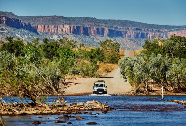 Ein Geländewagen fährt durch eine seichte Wasserstraße, umgeben von Buschland und mit Bergen in der Ferne, Pentecost River Crossing, Gibb River Road, Westaustralien © Tourism Western Australia