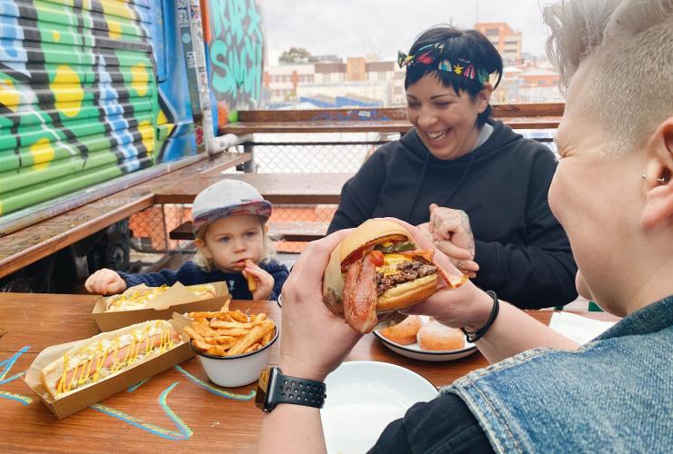 Eine Familie sitzt lachend an einem Tisch im Außenbereich neben einer farbenfroh bemalten Wand und isst Hamburger und Hot Dogs im Easey's, Collingwood, Victoria © Easey's
