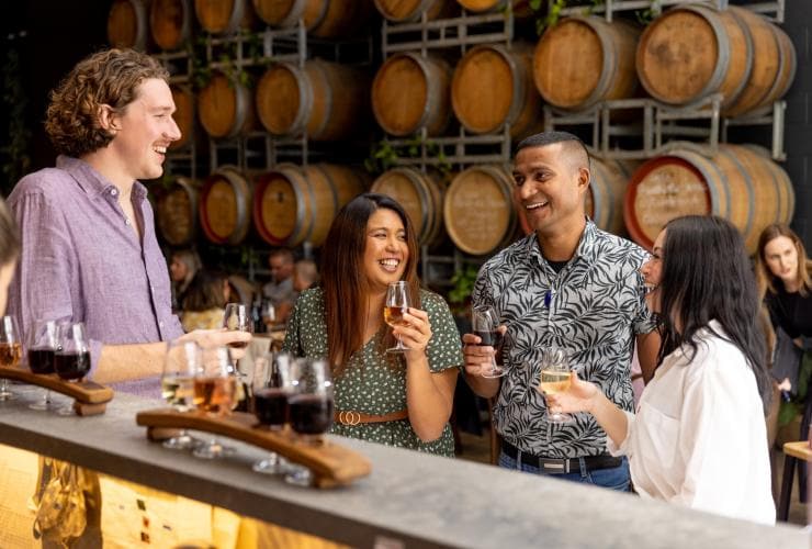 Einige Freunde genießen ein Glas Wein an einem Tisch vor einer Wand aus Fässern in der City Winery, Brisbane, Queensland © Tourism and Events Queensland