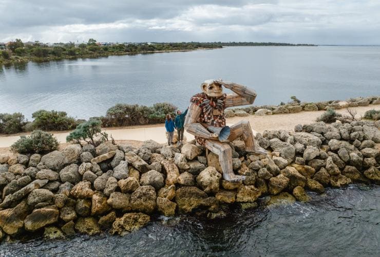 Zwei Menschen stehen neben der Skulptur eines Riesen, der auf einer Mauer im Meer sitzt, Seba's Song Giant, Giants of Mandurah, von Thomas Dambo, Mandurah, Westaustralien © Visit Mandurah