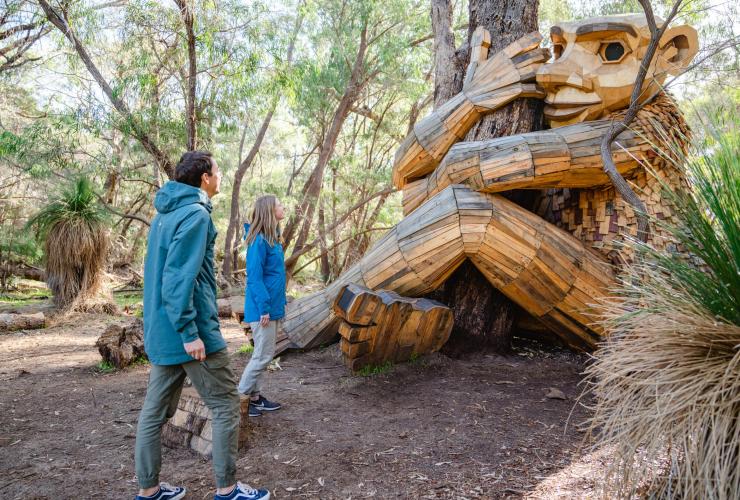 Zwei Menschen stehen neben der Skulptur eines Riesen, der einen Baum umarmt, Secret Giant Jyttes Hytte, Giants of Mandurah, von Thomas Dambo, Mandurah, Westaustralien © Visit Mandurah