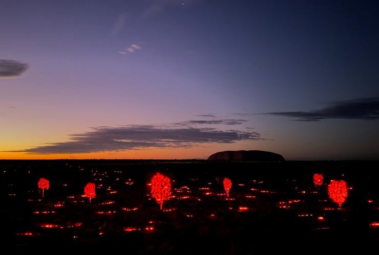 Eine Outback-Landschaft, die während der Morgendämmerung mit einer beleuchteten Kunstinstallation geschmückt ist, mit einer großen Felsformation in der Ferne bei Sunrise Journeys, Uluru-Kata Tjuta National Park, Northern Territory © Voyages Indigenous Tourism Australia