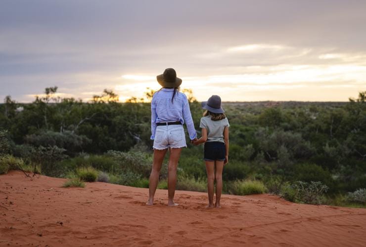 Mutter und Tochter halten sich an den Händen und stehen mit Blick auf das Buschland der Bullara Station auf dem roten Sand, Ningaloo, Westaustralien © Tourism Australia