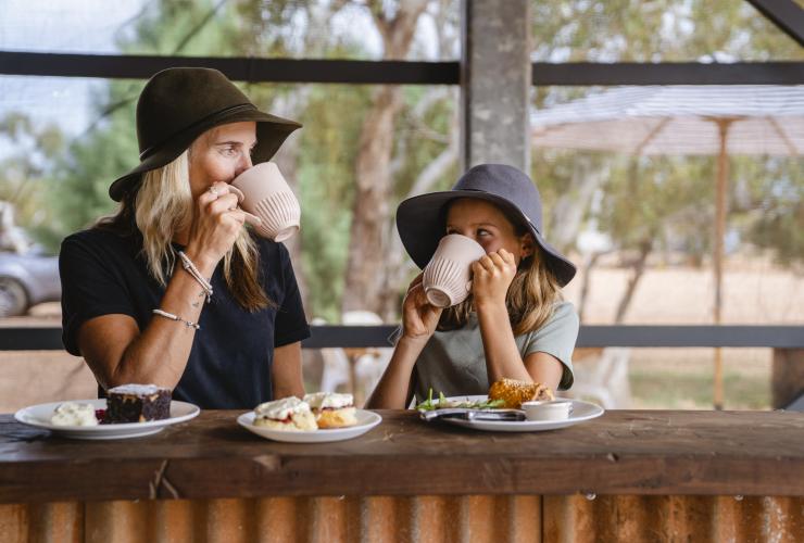 Mutter und Tochter essen vor der Kulisse des Buschlands hinter Bullara Station Gebäck und trinken aus Bechern, Ningaloo, Westaustralien © Tourism Australia