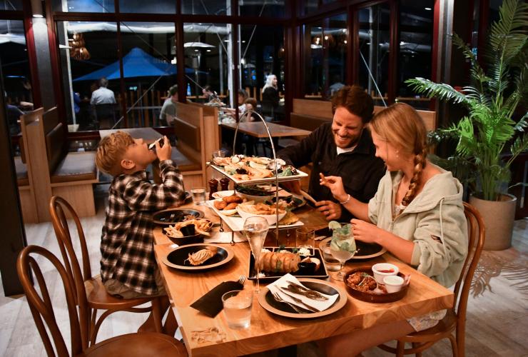 Eine Familie unterhält sich lachend in einem Restaurant an einem Tisch des Kingfisher Bay Resorts, der mit Essen gedeckt ist, K'gari, Queensland © Tourism and Events Queensland