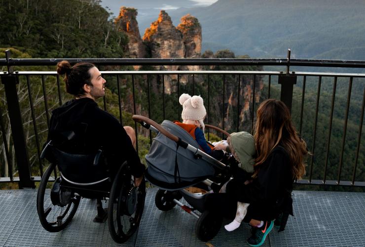 Eine Person im Rollstuhl mit Familie an einem Aussichtspunkt mit Blick auf drei Sandsteinsäulen, die sich aus einem weitläufigen Tal erheben, The Three Sisters, Blue Mountains, New South Wales © Tourism Australia