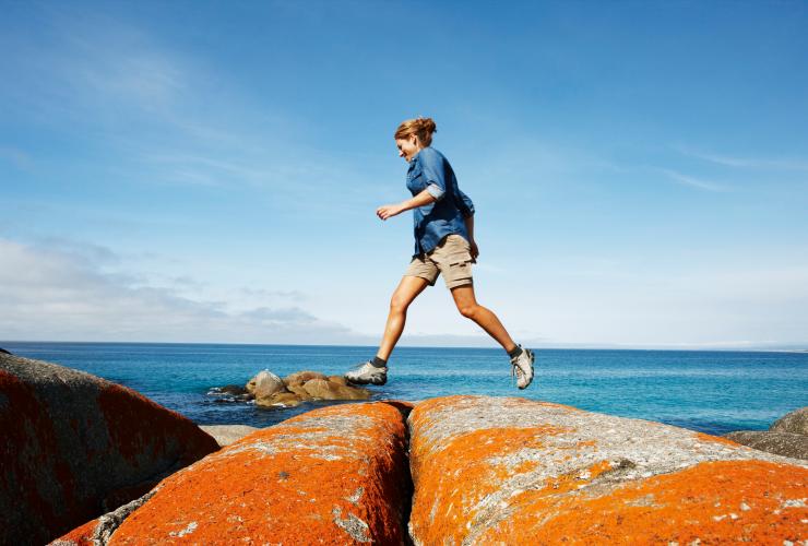 Eine Person hüpft über mit orangefarbenen Flechten bewachsene Felsen am Meer, Binalong Bay, Bay of Fires Conservation Area, Tasmanien © Tourism Australia
