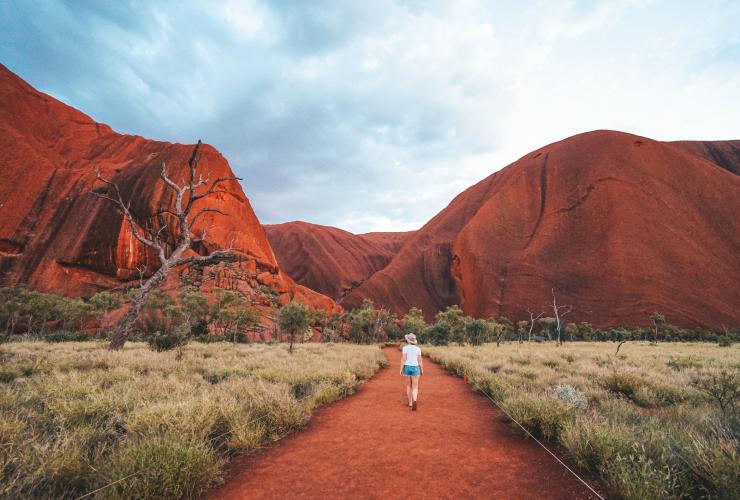 Eine Person, die auf einem Wanderweg an einem großen roten Felsenmonolithen entlanggeht, Uluṟu, Red Centre, Northern Territory © Tourism NT/Jackson Groves