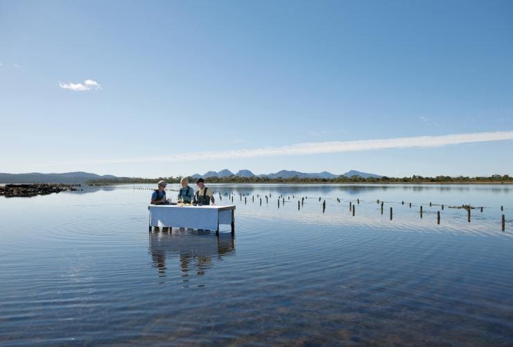 Zwei Personen stehen während des Austernknackens zum Abendessen mit einem Reiseführer am Wasser der Freycinet Marine Oyster Farm, Saffire Freycinet, Tasmanien © Tourism Tasmania