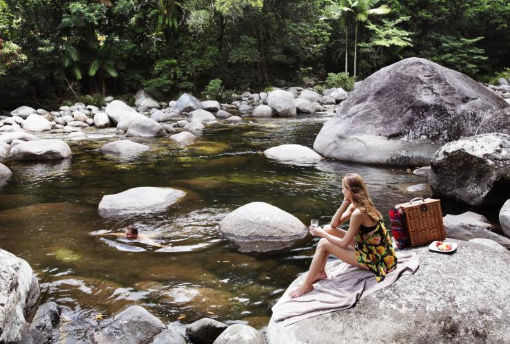 Eine Frau sitzt mit einem Glas Wein und einem Picknickkorb vor einem glatten Felsbrocken und beobachtet einen Mann, der im klaren Wasser des Mossman Gorge River in der Nähe der Silky Oaks Lodge schwimmt, Queensland © Luxury Lodges of Australia