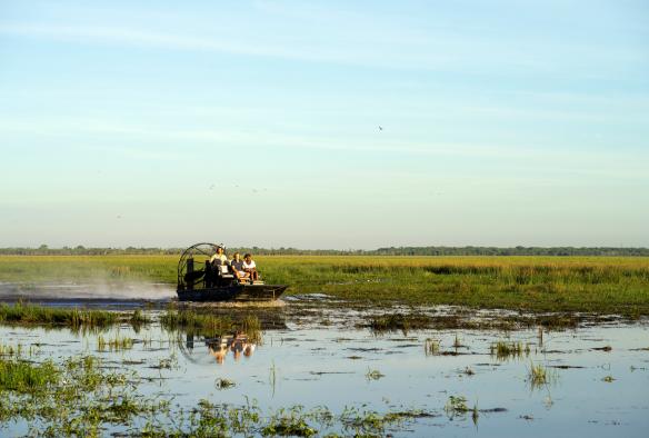Drei Personen in einem Luftkissenboot, das durch grüne Feuchtgebiete fährt, Bamurru Plains, Mary River, Northern Territory © Tourism NT/KWP!/Shaana McNaught