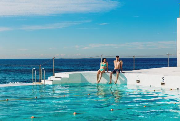 Ein Mann und eine Frau sitzen am Rand der Bondi Icebergs, die Füße im Wasser und das Meer hinter sich, Sydney, New South Wales © Destination NSW