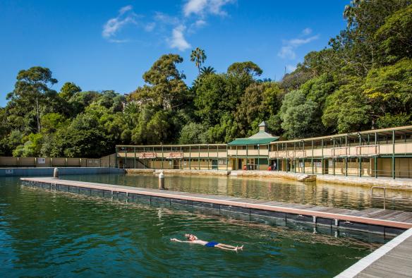 Eine Person schwimmt im blaugrünen Wasser der Dawn Fraser Baths mit den historischen Mauern des von Bäumen umgebenen Gebäudes im Hintergrund, Balmain, Sydney, New South Wales © Destination NSW