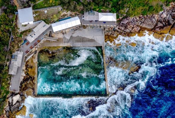 Luftaufnahme des Ozean-Pools „Wylie's Baths“ mit schwimmenden Menschen und Wellen, die über die Küstenfelsen rund um den Pool schlagen, Coogee, New South Wales © Destination NSW