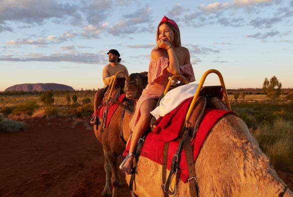 Ein Mann und eine Frau blicken auf die Landschaft des Uluru-Kata Tjuta National Park mit dem Uluru im Hintergrund, während sie mit Uluru Camel Tours auf Kamelen reiten, Northern Territory © Tourism NT/Matt Cherubino