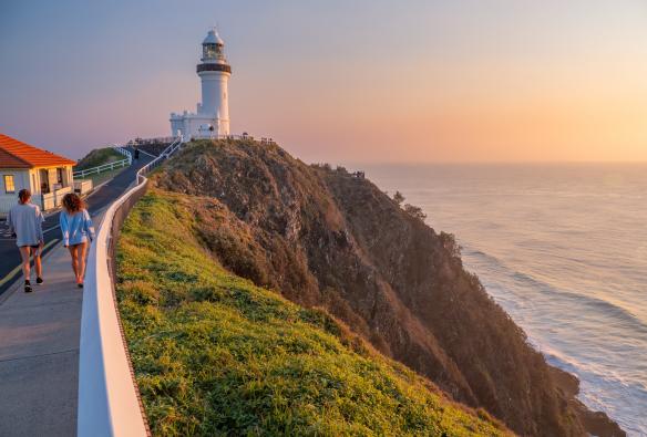 Zwei Menschen gehen auf einem Weg zum Cape Byron Bay Lighthouse, neben ihnen ein grasbewachsener Hügel, der zum Meer hin abfällt, Byron Bay, New South Wales © Tourism Australia