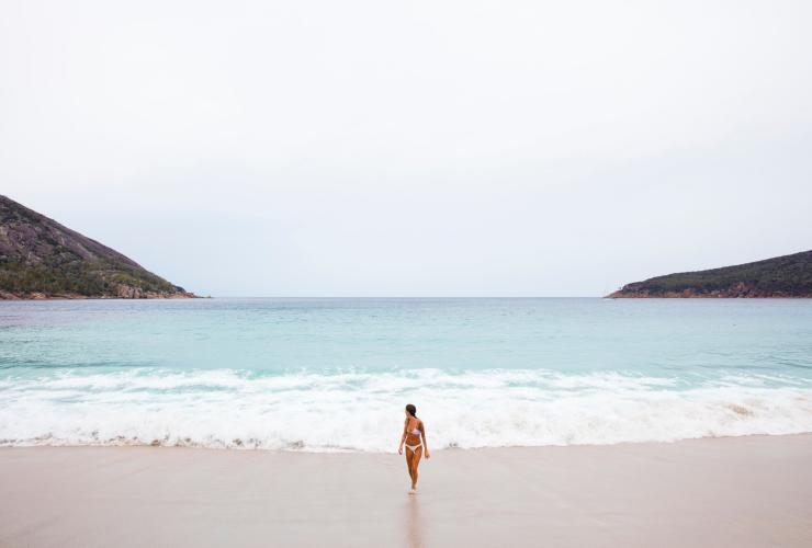 Eine Frau kommt aus dem Meer und geht auf dem weißen Sand am Wineglass Bay Beach, Freycinet National Park, Tasmanien © Sean Scott