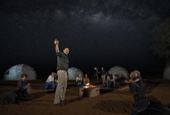 Ein Tour Guide zeigt auf den mit Sternen übersäten Nachthimmel, während die Gäste im Kreis um ein Lagerfeuer sitzen und zuhören, Kuppelzelte stehen im Hintergrund, Earth Sanctuary, East MacDonnell Ranges, Northern Territory © Tourism Australia