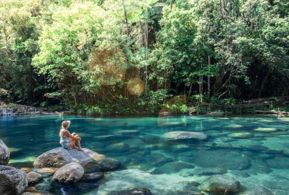 Eine Frau sitzt auf einem großen Felsbrocken in einem Naturpool mit klarem blauem Wasser, der von Bäumen umgeben ist, Mossman Gorge, Mossman, Queensland © Tourism and Events Queensland