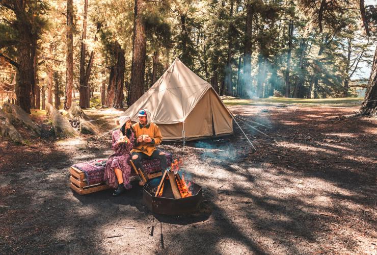 Ein Paar sitzt mit Bechern in den Händen vor einem Lagerfeuer vor einem Zelt und Bäumen im Mount Crawford Forest, Adelaide Hills, Südaustralien © We Live We Explore