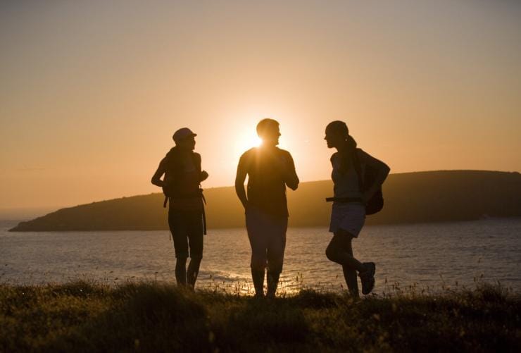 Drei Personen wandern mit Rucksäcken, während die Sonne an Kings Head über dem Meer untergeht, Fleurieu Peninsula, Südaustralien © South Australian Tourism Commission/Peter Fisher