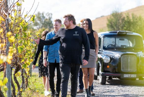 A tour guide from Van Du Vin leads a small tour group past some green grapevines. Behind them is the black, old English-style taxi cab used to transport guests on the Van Du Vin tours, Canberra, Australian Capital Territory © Tourism Australia 