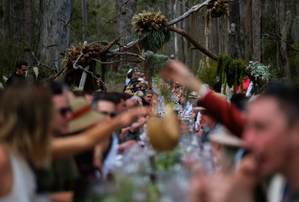 A striking scene shows diners at a very long dining table covered in a white table cloth, situated on a bush track surrounded by gum trees.