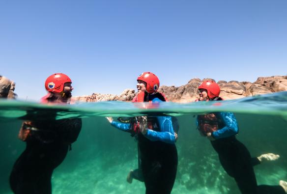 A semi-submerged shot shows a group of people wearing wetsuits, life vests and helmets, floating in turquoise ocean water with Margaret River Adventure Co, Margaret River, Western Australia © Tourism Australia