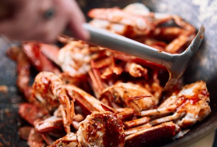 A close up photo shows a tumble of crabs being wok fried with chilli in a large, industrial sized wok. The chef’s hand is seen stirring the crab with a metal spatula. The crab looks fresh and succulent, Shuck & Shell Seafood Festival, Barrington Coast, New South Wales © Shuck & Shell Seafood Festival