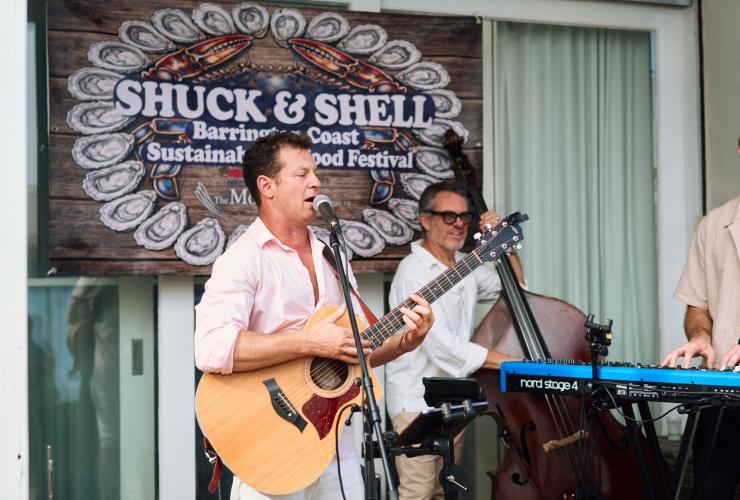In front of a large banner showing the branding for the Shuck & Shell Seafood Festival are three musicians, playing instruments. They’re laidback and happy as they play live music to the festival crowd, Barrington Coast, New South Wales © Shuck & Shell Seafood Festival