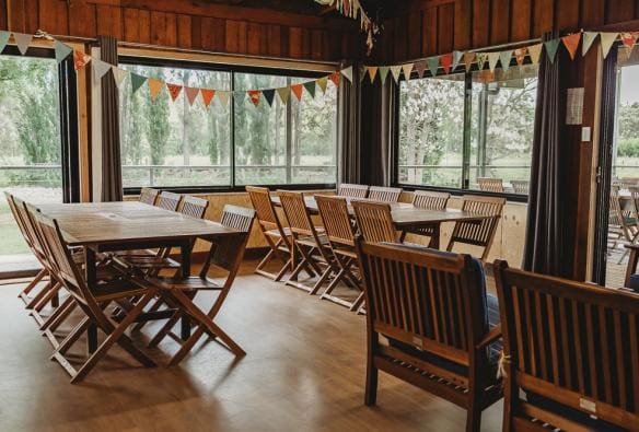 Taken from inside a wooden hall, there is a collection of wooden tables and chairs, arranged neatly, ready to accommodate several groups of people at The Orchard Lodge, Barrington Coast, New South Wales © The Orchard Lodge