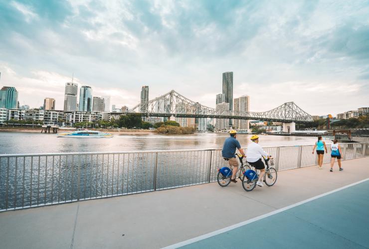 People walking and cycling along a pathway beside Brisbane River with the city skyline in the distance along the River Walk, Brisbane, Queensland © Tourism and Events Queensland