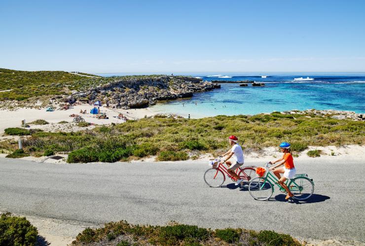 Two people cycling on coloured bikes along a road beside a bay with white sand and clear blue water at Little Salmon Bay, Rottnest Island, Western Australia © Tourism Western Australia