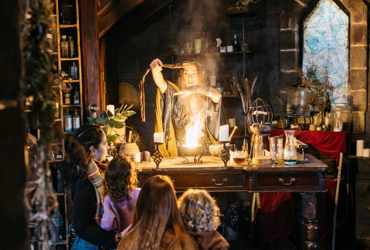 Parents and children watching as an actor dressed in a fairytale costume prepares a fiery potion surrounded by props at Kryal Castle, Ballarat, Victoria © Tourism Australia/Visit Victoria