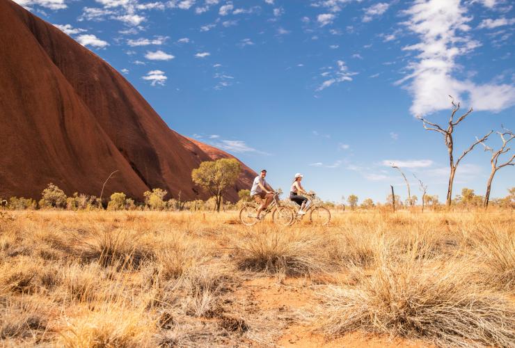 Two people cycling past a towering red rock formation, Uluṟu-Kata Tjuṯa National Park, Uluru, Northern Territory © Tourism NT/KWP!