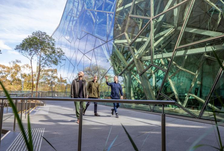 Two people walking past an architectural building with a tour guide, Localing Tours, Melbourne, Victoria © Tourism Australia