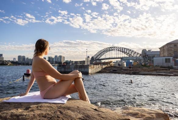 A woman sitting on a sandstone rock looking past Marrinawi Cove to the Sydney Harbour Bridge while people swim below her, Barangaroo Reserve, Sydney, New South Wales © Images for Business
