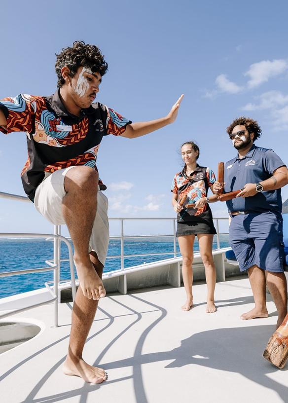 Aboriginal tour guides dancing and playing traditional instruments on a boat in the ocean.