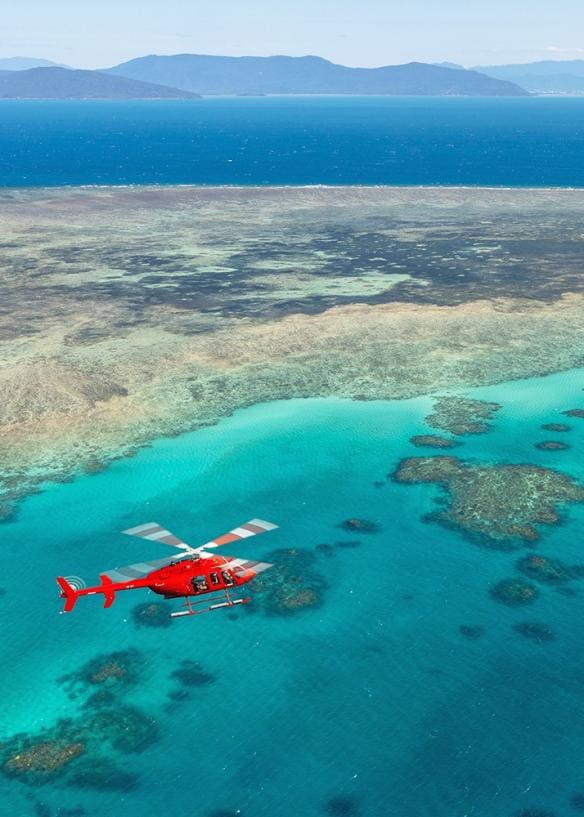 A red Nautilus Aviation helicopter flying above coral reefs towards a lush island.