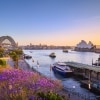 A city harbour during sunset with boats, a tall arched bridge and jacarandas in the foreground, Sydney Harbour, Sydney, NSW © Destination NSW