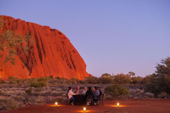 Group gathered around an outdoor dining table with Uluru in the background, Uluru-Kata Tjuta National Park, Northern Territory© Tourism Australia
