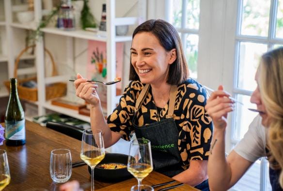 Two people are sitting down to eat at a shared table. They look happy, and are chatting to others not pictured, as they enjoy the meal they prepared as part of the Harvest Experience cooking class. Sitting on the table along with their food are glasses of local wine, Harvest Experience, Geelong and the Bellarine, Victoria © Harvest Experience