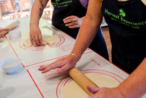 Two people stand at a kitchen-style bench, taking part in the cooking class at Harvest Experience. They are both laughing as they work on rolling out dough with rolling pins. They are both wearing aprons with ‘Harvest Experience’ embroidered on the front, Geelong and the Bellarine, Victoria © Harvest Experience