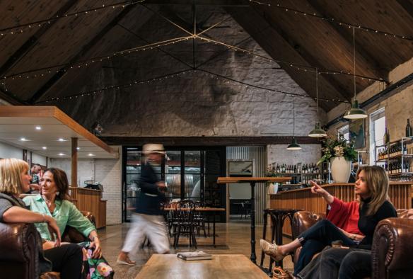 Inside a cosy winery barrel hall, with a pitched high roof, a tour group sits on couches at a low table. Behind a tour guide walks around fetching tastings for the group to sample, Epic Taste Experiences, Geelong and the Bellarine, Victoria © Epic Taste Experiences
