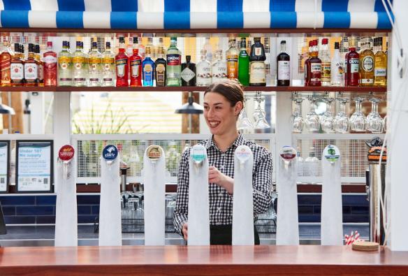 A smiling bar staffer stands behind beer taps, pulling a beer. Above her is a row of alcohol used to make cocktails, Portarlington Grand Hotel, Geelong and the Bellarine, Victoria © Tourism Australia