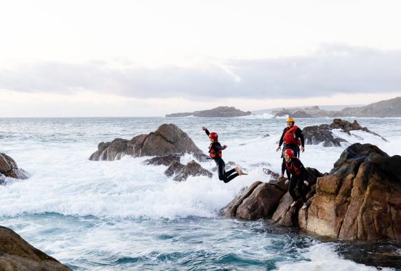 A person wearing a wetsuit, life vest and helmet is seen jumping from a rock into pristine ocean water below on a tour with Margaret River Adventure Co, Margaret River, Western Australia © Tourism Australia