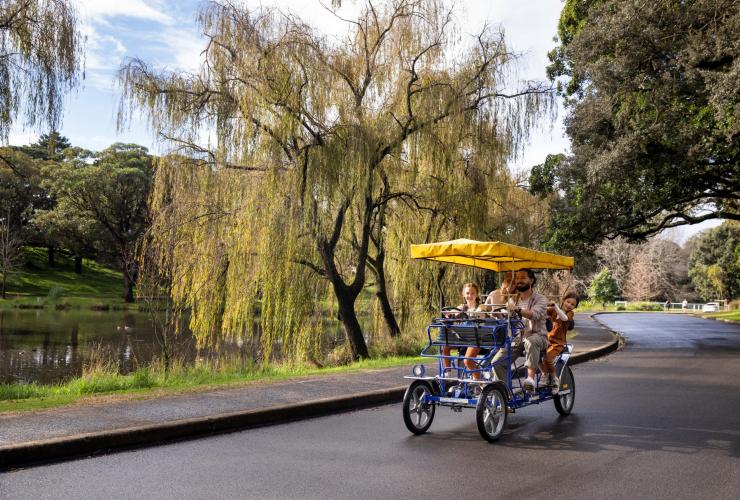 A family riding on a four-wheeled bike with an umbrella past a pond through the greenery of Centennial Park Cycles, Centennial Park, New South Wales © Destination NSW