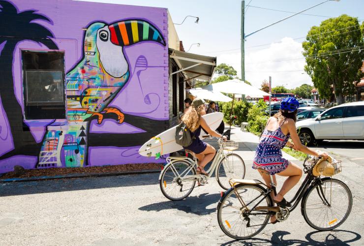 Two people cycling past a colourful mural of a toucan, one holding a surfboard in Byron Bay, New South Wales © Destination NSW/James Horan